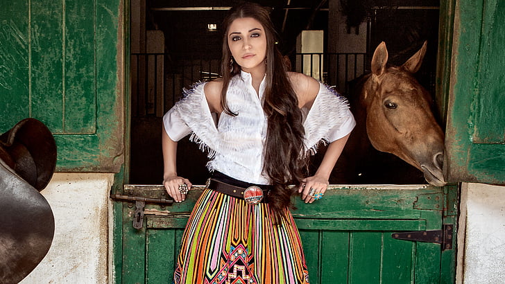 woman in white button-up top in front of brown horse inside stable