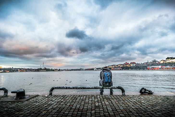 person in gray jacket and blue backpack sitting on black steel tube bench beside body of water under white clouds