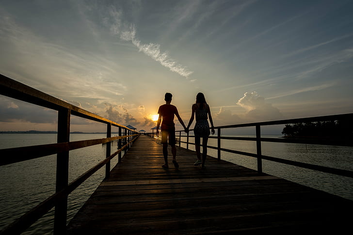 silhouette of man and woman holding hands walking on brown wooden dock during golden hour