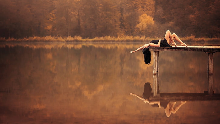 women's black dress, woman lying on wooden dock near body of water during daytime