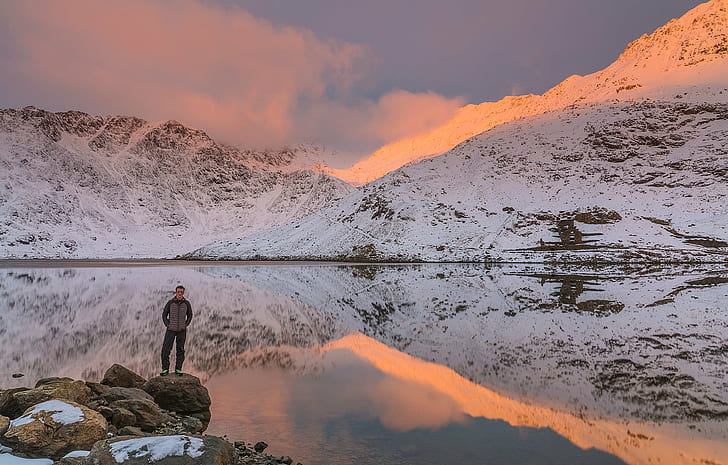 photo of man in jacket standing on big rocks beside body of water with white mountain during daytime, snowdonia, snowdonia