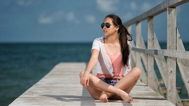 black haired woman in sunglasses sitting on dock beside body of water