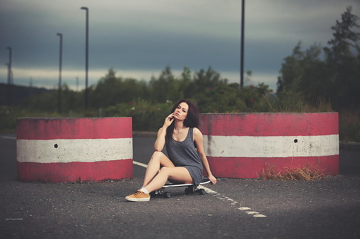 woman in grey sleeveless dress sitting on skateboard posing with one hand on face