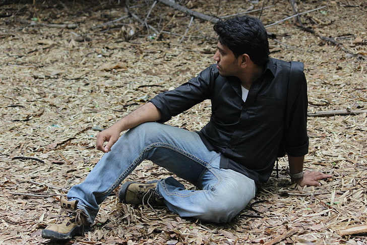 man in black dress shirt and blue jeans sitting on ground, waiting