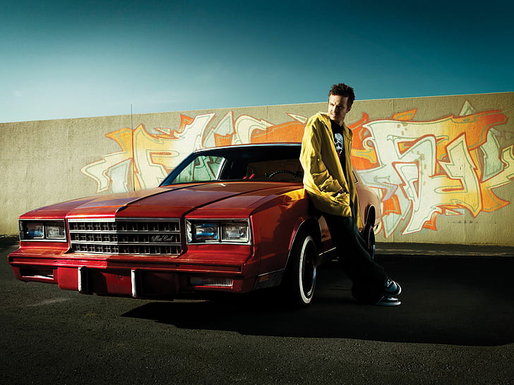 man wearing yellow jacket leaning red car in front of wall artwork