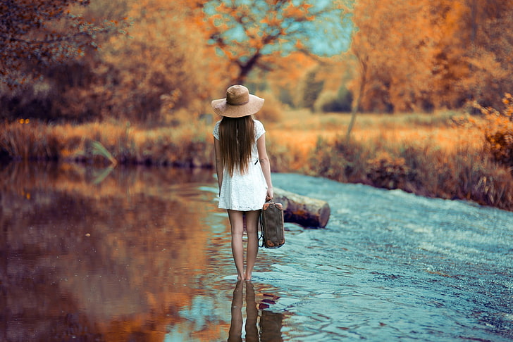 women's white cap-sleeved mini dress, woman walking on shallow body of water carrying suitcase under shad of tree at daytime