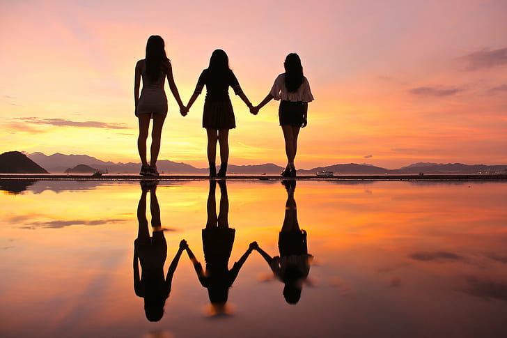 silhouettes of  women staining near a water under orange sky