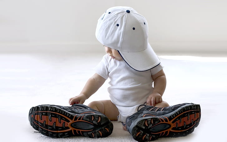 Wearing a pair of big shoes cute baby, baby's white onesie; white cap; black lace up hiking shoes;