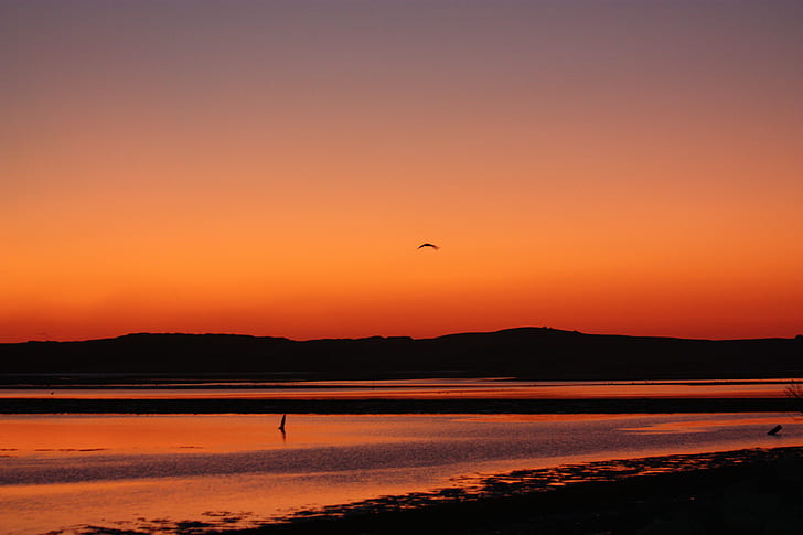 silhouette of hill with orange sky mirrored on body of water