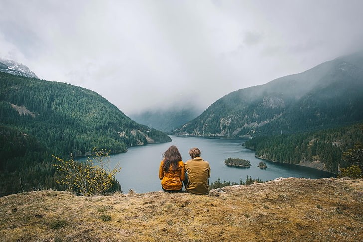 two person sitting on cliff near lake surrounded by mountains