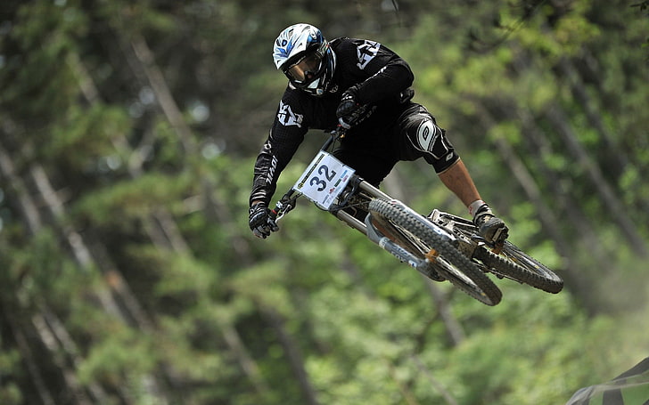 mountain bike racer wearing black-and-white long-sleeved shirt and pants and white full-face helmet soaring on air downhill during daytime