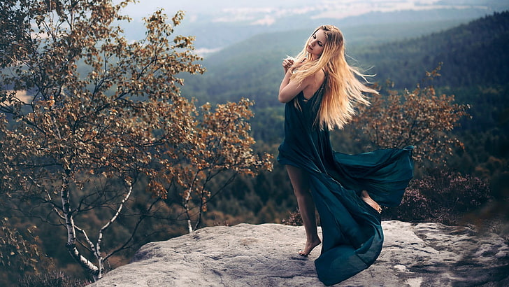 women's blue sleeveless slit dress, woman wearing teal sleeveless dress posing with both of her hands on her heart while she close her eyes on grey cliff surrounded with brown trees