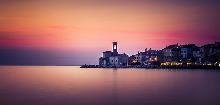 calm body of water with lighted buildings under pink and orange sky, piran, piran